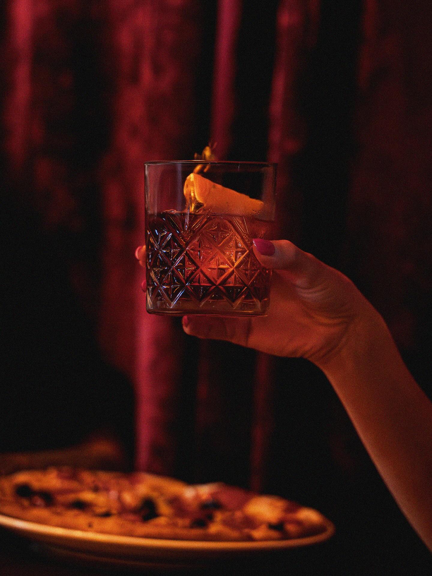 A cut-glass negroni with an orange twist held up against the Gemm's red velvet, a pizza below