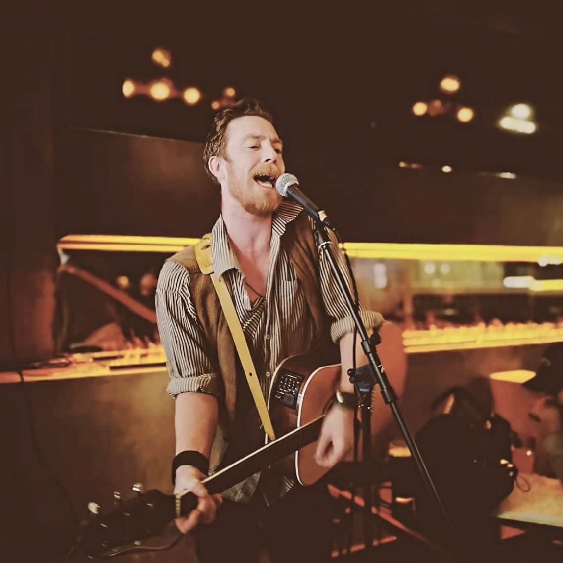 A guitarist singing into the mic under the lights at the back of the room