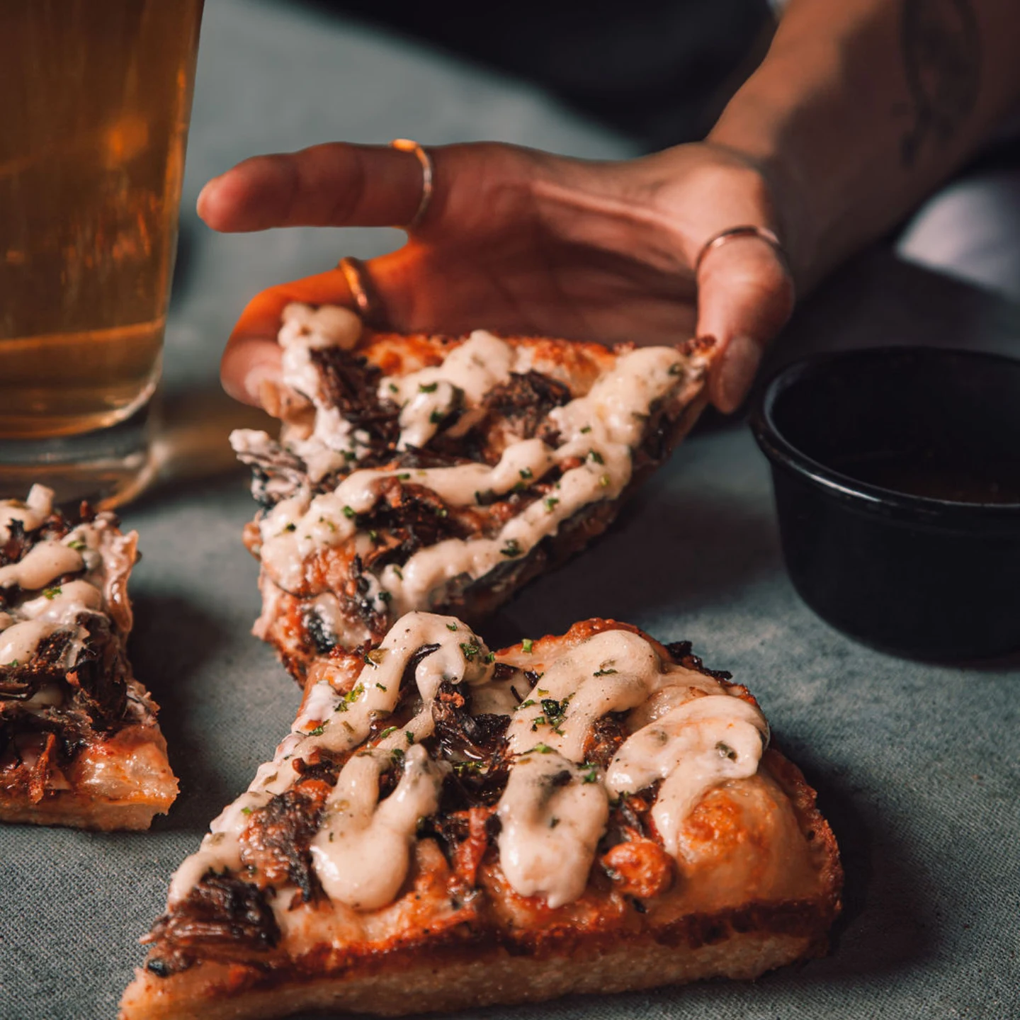 A beef dip slice being lifted, drizzled with garlic cream, with au jus and a pint alongside